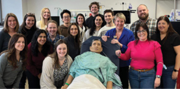 a diverse group of medical students posing with a simulator model on a gurney
