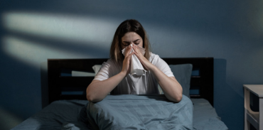 a women blowing her nose into a tissue while sitting up in bed